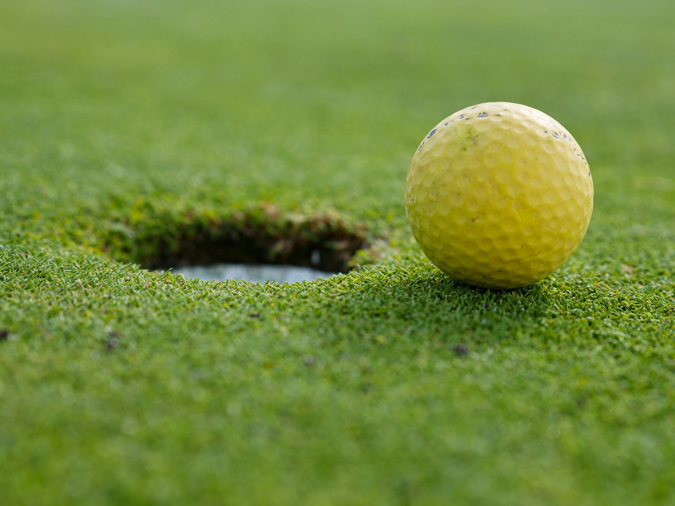 A yellow golf ball close to the hole on a grassy golf course.